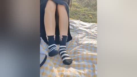 Video of a person with fair skin, wearing a navy dress and navy socks with white stripes, sitting on a yellow and white checkered blanket in a grassy outdoor setting.