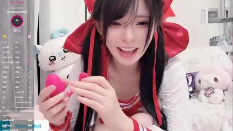 A video of an Asian woman with long black hair in a red bow, smiling while holding a pink vibrator, surrounded by plush toys and a video game screen.