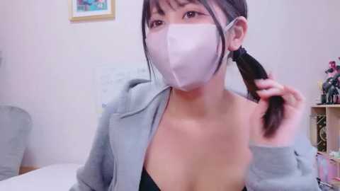 A video of an East Asian woman with long black hair in a ponytail, wearing a grey hoodie and a face mask. She stands in a room with a desk, framed picture, and plants in the background.