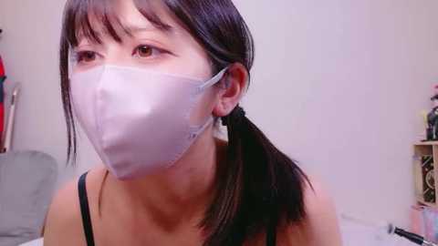 A video of an East Asian woman with long black hair in pigtails, wearing a white face mask and black spaghetti strap top, indoors with a grey chair and shelves in the background.
