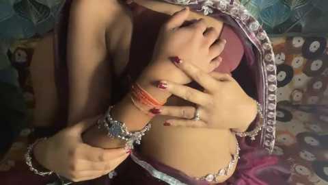 Video of a woman with fair skin, wearing a traditional, ornate, purple sari, adorned with silver and red jewelry. Her hands gently touch her chest, revealing a hint of cleavage. Background features a colorful, patterned fabric.