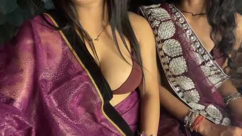 Video of two women, one in a maroon bra, the other in a purple sari with silver embroidery, seated closely together, with a black shoulder strap between them.