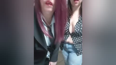 Video of two women, one with long, maroon hair, wearing a black jacket, the other with medium-length brown hair, a polka-dot top, and light blue jeans.