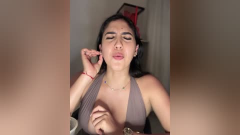 Video of a young woman with olive skin, dark hair, and closed eyes, wearing a halter top, touching her lips, set in a dimly lit room with a blurred background.