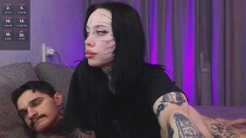 Video of a tattooed, pale-skinned woman with dark hair, wearing a black shirt, holding a man's head, in a dimly lit bedroom with purple lighting.