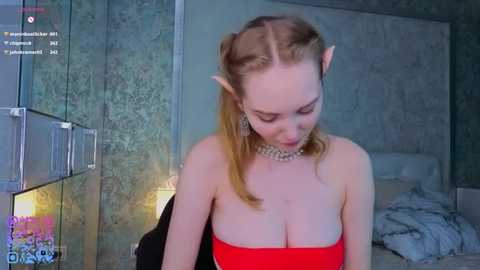 Video of a fair-skinned, blonde-haired woman with pointed elf ears, wearing a strapless red dress, in a dimly lit room with teal walls.