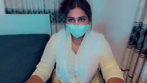 Video of a South Asian woman with medium skin tone, wearing a light yellow kurta, light blue mask, and dark hair, sitting on a bed with blue sheets in a modestly decorated room.
