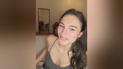 A young woman with long, dark hair, fair skin, and a slender physique, wearing a gray spaghetti-strap top, smiles while leaning against a beige wall. Behind her, a mirror reflects part of the room, including a wooden table.