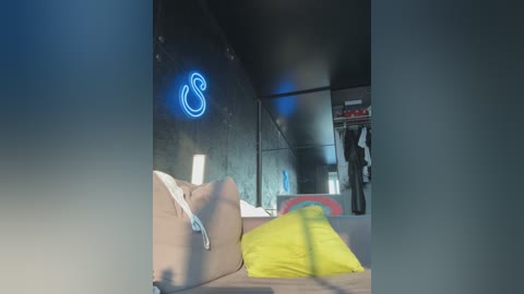 Video of a modern, dimly lit bedroom with a neon \"S\" sign on the wall, a person in a white shirt lying on a bed, and a yellow pillow in the foreground.