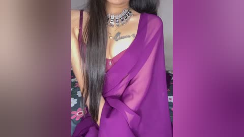 Video of a woman with long black hair, wearing a sheer purple sari, silver necklace, and revealing a tattoo on her chest, in a room with a floral-patterned bedspread.