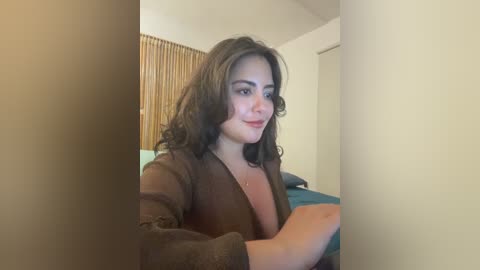 A video of a young woman with medium brown skin, long dark hair, and a slim figure, wearing a brown cardigan, standing in a beige room with wooden blinds and a blue bedsheet.