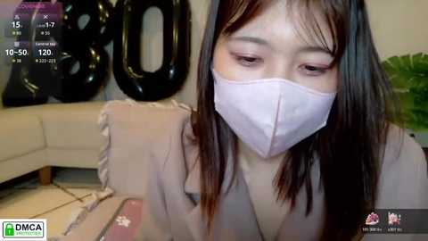 Video of an Asian woman with long black hair, wearing a pink face mask, sitting on a beige sofa with a black \"200\" balloon in the background.