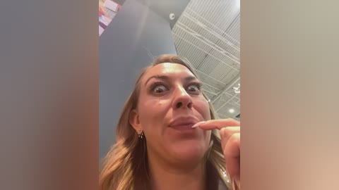 Video of a smiling, light-skinned woman with long, wavy brown hair, wearing a gold earring, and touching her lips with her index finger, taken from a low angle inside a building with a high ceiling and white panels.