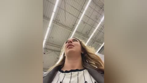Video of a young woman with fair skin and long blonde hair, wearing a white and black striped blouse and a gray blazer, standing inside a modern industrial building with a high ceiling and exposed metal beams.