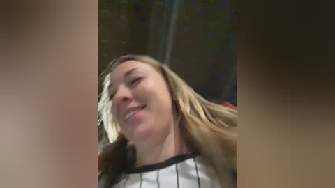 Video of a smiling woman with long blonde hair, wearing a black and white striped shirt, in a blurred outdoor setting with greenery.