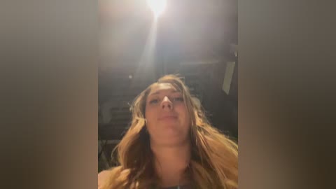 Video of a young woman with long, wavy, blonde hair, gazing upward at a bright light source. She has a fair complexion and appears relaxed. The background is dimly lit, suggesting an indoor setting.