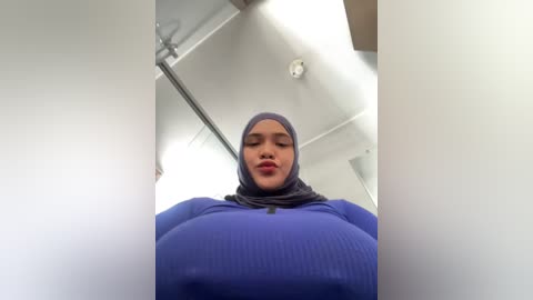Video of a young woman with medium skin tone, wearing a tight blue ribbed top, gray hijab, and no makeup, standing in a modern, brightly-lit room with white walls and a glass door.