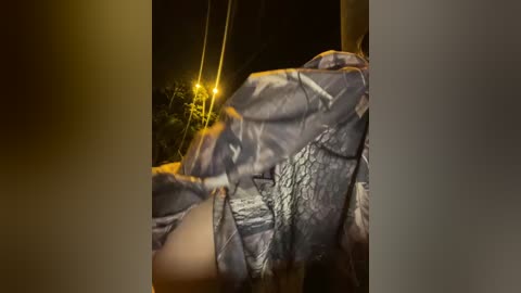 A nighttime video captures a person wrapped in a blue and white camouflage blanket, with a yellow streetlight illuminating their partially visible face.