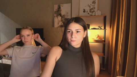 Video of two young women in a cozy room. One has long dark hair, wearing a black sleeveless top, standing in the foreground. The other, blonde, with short hair, wearing a white T-shirt, adjusts her hair in the background.