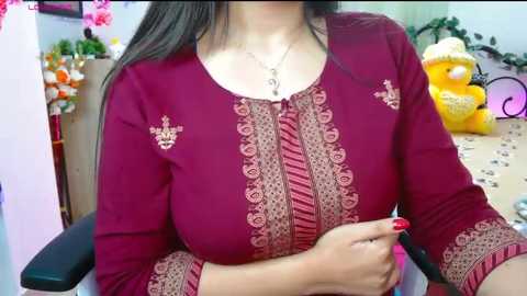 Video of a woman with long dark hair, wearing a deep red, long-sleeved blouse with intricate embroidery, seated in a room with floral decorations, holding a pen.
