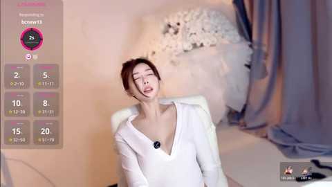 A video shows an Asian woman with dark hair, wearing a white blouse, and a black necklace, lying on a white couch in a dimly lit room.