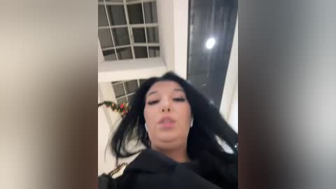 A video of a woman with long black hair and full lips, wearing a black jacket, taken from a low angle, showing a modern, well-lit room with a glass block wall.