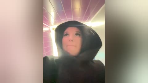 Video of a person in a black hooded sweatshirt, standing in a pink-tiled hallway with white ceiling tiles, wearing sunglasses.