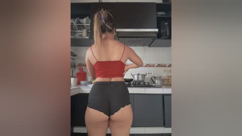 Video of a woman with a ponytail, wearing a red tank top and black lace-trimmed shorts, cooking in a modern kitchen with white tiles and stainless steel appliances.