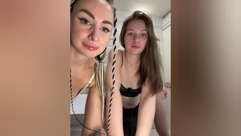 Video of two young women, one with light skin and braided hair, the other with fair skin and long brown hair, both wearing black lingerie, in a modern indoor setting.