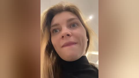 A close-up video of a Caucasian woman with light skin, brown hair, and blue eyes, wearing a black turtleneck. The background is blurred, showing beige walls and a ceiling.