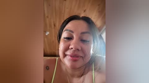 Video of a smiling, light-skinned Asian woman with dark hair, wearing a green top, standing in a wooden-paneled room.