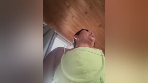 A low-angle video captures a woman with medium-length brown hair and fair skin, wearing a lime green top, gazing upward. The background features a wooden ceiling and a partially visible door.