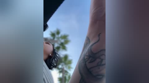 Video of a person with short black hair, seen from a low angle, standing between two trees with a clear blue sky background. The person's right arm has a detailed, black ink tattoo.