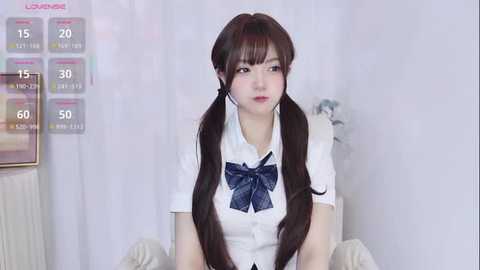 Video of an Asian woman with long, straight brown hair in twin pigtails, wearing a white schoolgirl uniform with a blue bow. She sits in a white chair against a white backdrop.