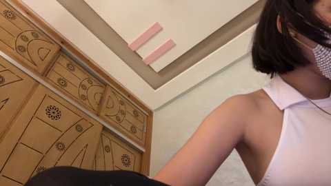 A video of a woman with short black hair, wearing a white sleeveless top, standing next to a wooden panel with abstract designs and pink vertical strips.