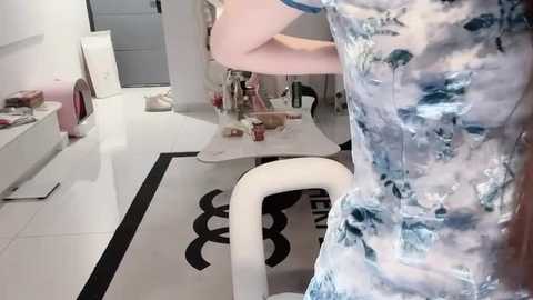 A video of a person wearing a white floral dress with blue accents, standing in a modern, minimalist room with white walls, black and white tiled floors, and a white chair.