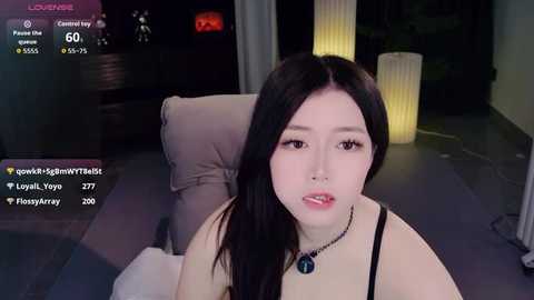 Video of an East Asian woman with long black hair, wearing a black top, sitting on a beige couch, indoors at night.