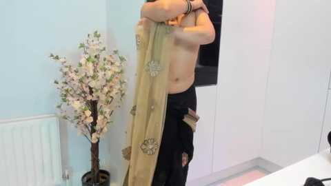 A video shows a light-skinned person, topless, holding a beige sari with intricate embroidery, standing in a minimalist room with white walls and a tall floral arrangement.