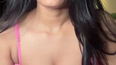 Video of a woman with long black hair, wearing a pink bra, showing her bare shoulders and upper chest. The background is blurred, indoor setting.