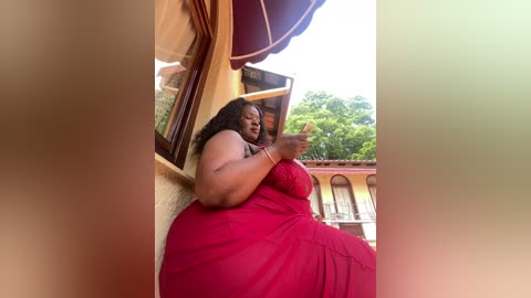 Video of a plus-sized Black woman in a red dress, sitting on a balcony railing, gesturing with her hands. Background shows a yellow building with arched windows and green trees.