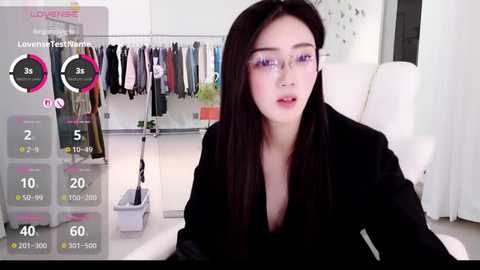 A video of a young Asian woman with long black hair, wearing glasses, sitting in a modern room with a white chair, a hanging clothes rack, and a digital screen displaying stats.