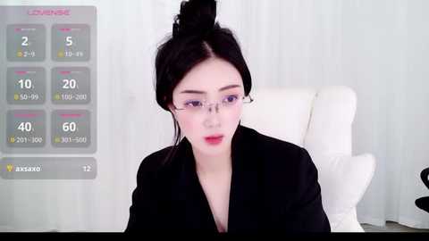 A video of an East Asian woman with fair skin, dark hair in a bun, wearing glasses, a black blazer, and red lipstick, sitting in a white chair, with a digital health app interface overlaid.