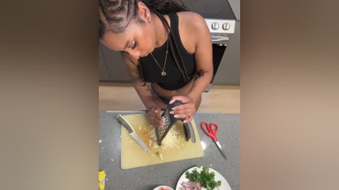 Video of a Black woman with braided hair, wearing a black crop top, slicing a cucumber on a kitchen counter.
