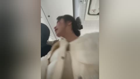 A blurred video of a person with short dark hair, wearing a beige coat, sitting in an airplane bathroom. The background shows a white-tiled wall and a towel holder.