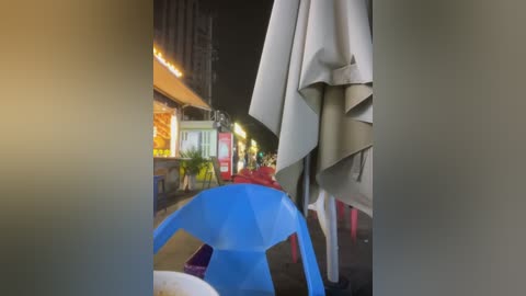 A nighttime video of a wet street scene with a blue plastic chair, a beige umbrella, and blurred pedestrians under warm street lights.