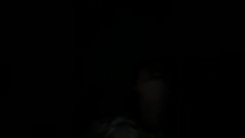 A dimly lit, grainy video shows a woman's silhouette with shoulder-length hair, partially obscured by shadows, against a pitch-black background. The image is slightly out of focus, adding to the mystery.