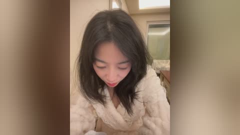 A video of an Asian woman with straight black hair, wearing a fluffy white robe, bending over, looking down, in a beige-tiled bathroom with a mirror and countertop.