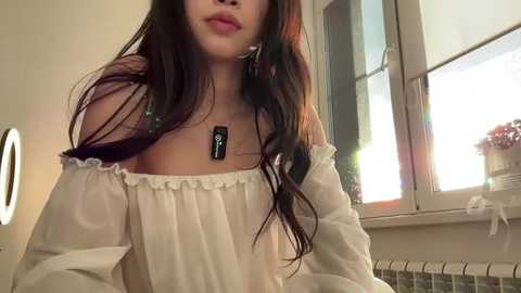 Video of an Asian woman with long, dark hair, wearing an off-the-shoulder white blouse, standing near a window with sunlight streaming in, holding a smartphone.