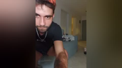 Video of a bearded man in a black t-shirt, leaning forward in a dimly lit, modern apartment with light-colored walls, a blue couch, and white doors.