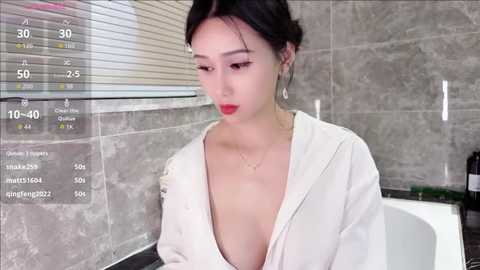 A video of an East Asian woman with fair skin, dark hair, and red lipstick, wearing a white robe, sitting in a modern bathroom with gray tiled walls and a bathtub.